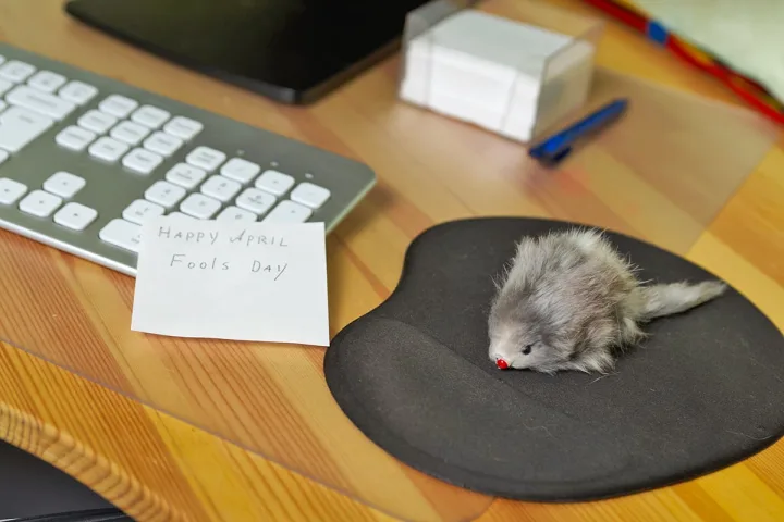 A desk with a toy mouse on a mouse pad and a handwritten 'Happy April Fools Day' sticky note beside a keyboard.