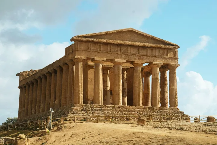 Ancient Greek-style stone temple with large columns under a partly cloudy sky.