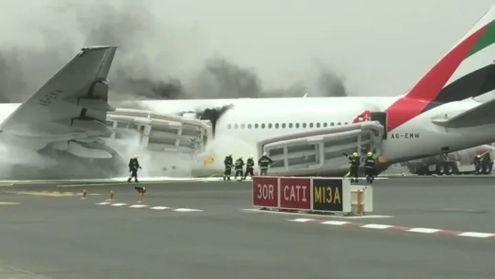 Firefighters on runway battling flames consuming an Emirates wide-body aircraft with heavy black smoke.