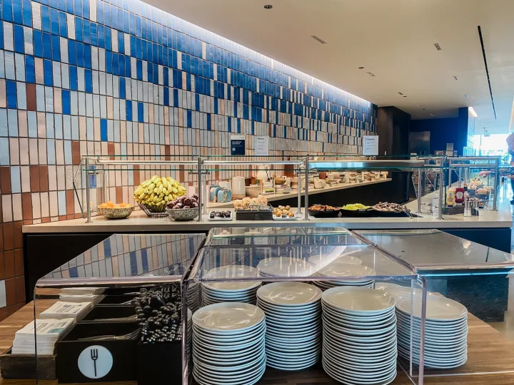 Well-stocked airport lounge buffet food station with stacked plates and tiled blue wall.