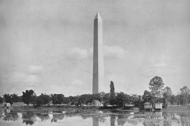 Black-and-white historical photo of the Washington Monument reflected in a pond, surrounded by trees.
