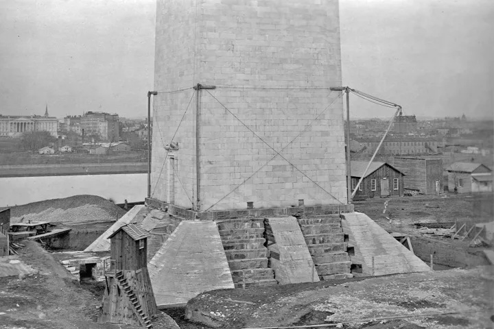 Historical black-and-white photo of the Washington Monument base under construction with scaffolding and cranes.