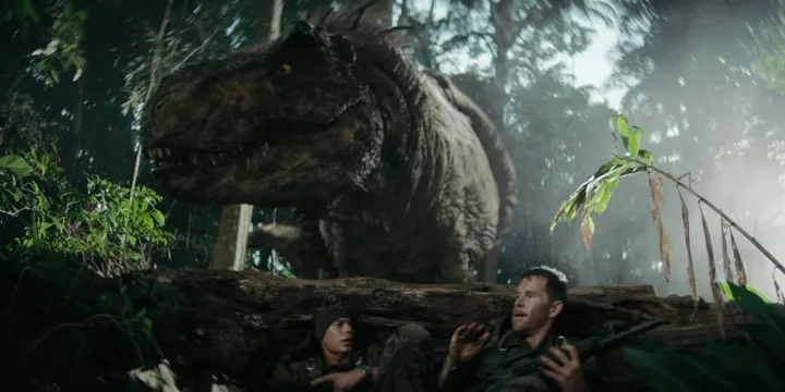 Two people hide behind a fallen log as a massive T-Rex looms over them in a jungle.