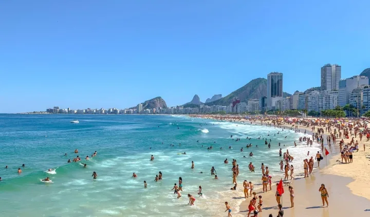 Crowded Copacabana beach in Rio de Janeiro with turquoise water, city skyline, and mountains.