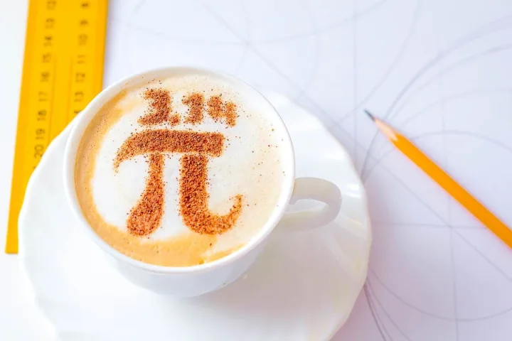 Latte in white cup with pi symbol drawn in cinnamon on foam, beside a ruler and pencil.