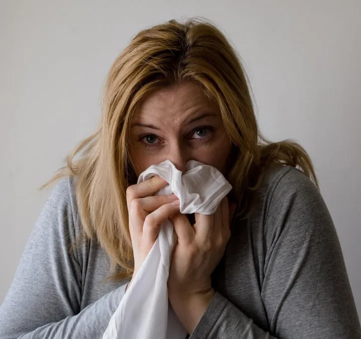 Blonde woman holding a tissue to her nose, looking ill against a plain background.