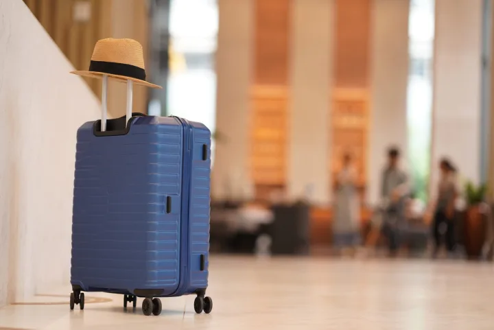 Blue hard-shell rolling suitcase with straw hat in a bright hotel lobby.