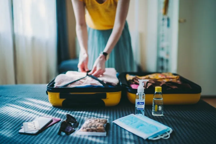 Woman packing a yellow open suitcase on a bed with travel items and a face mask nearby.
