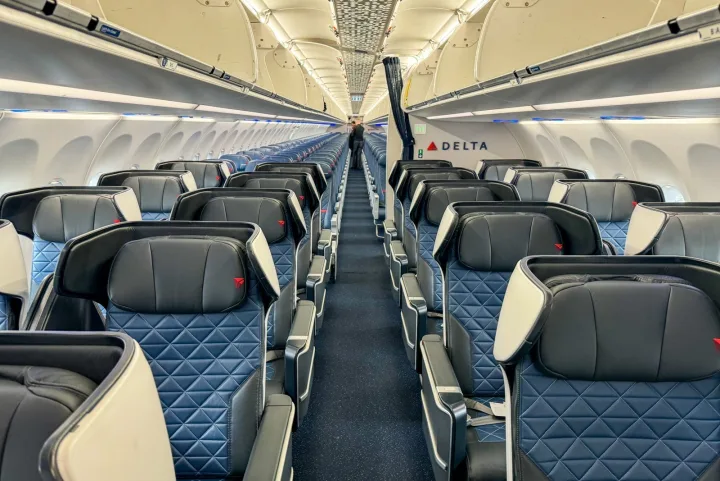 Wide empty Delta first class cabin interior with blue quilted seats and an attendant at the back.