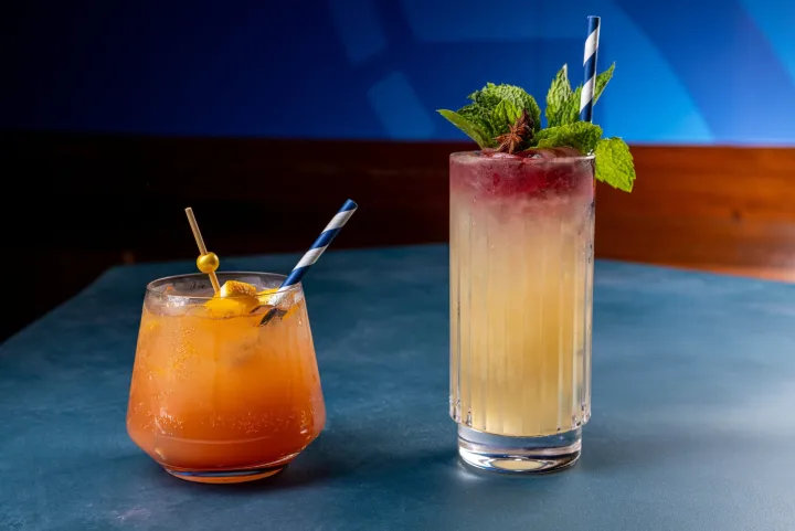 Two colorful cocktails with straws and garnishes on a blue table in a dim bar setting.