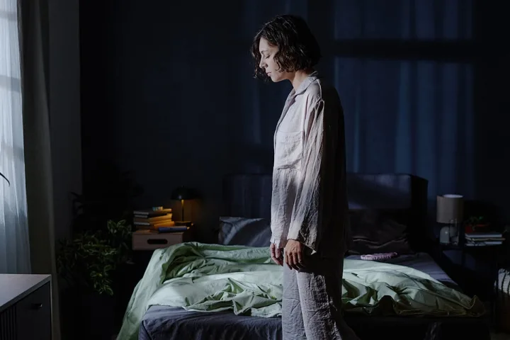 Woman in pajamas standing beside a bed in a dark bedroom, eyes downcast, dazed expression.