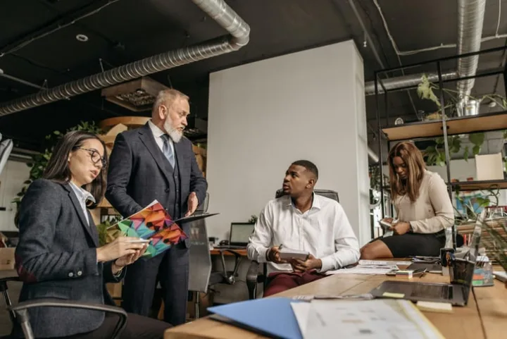 Senior executive standing over younger colleagues in a modern office, reviewing documents together.