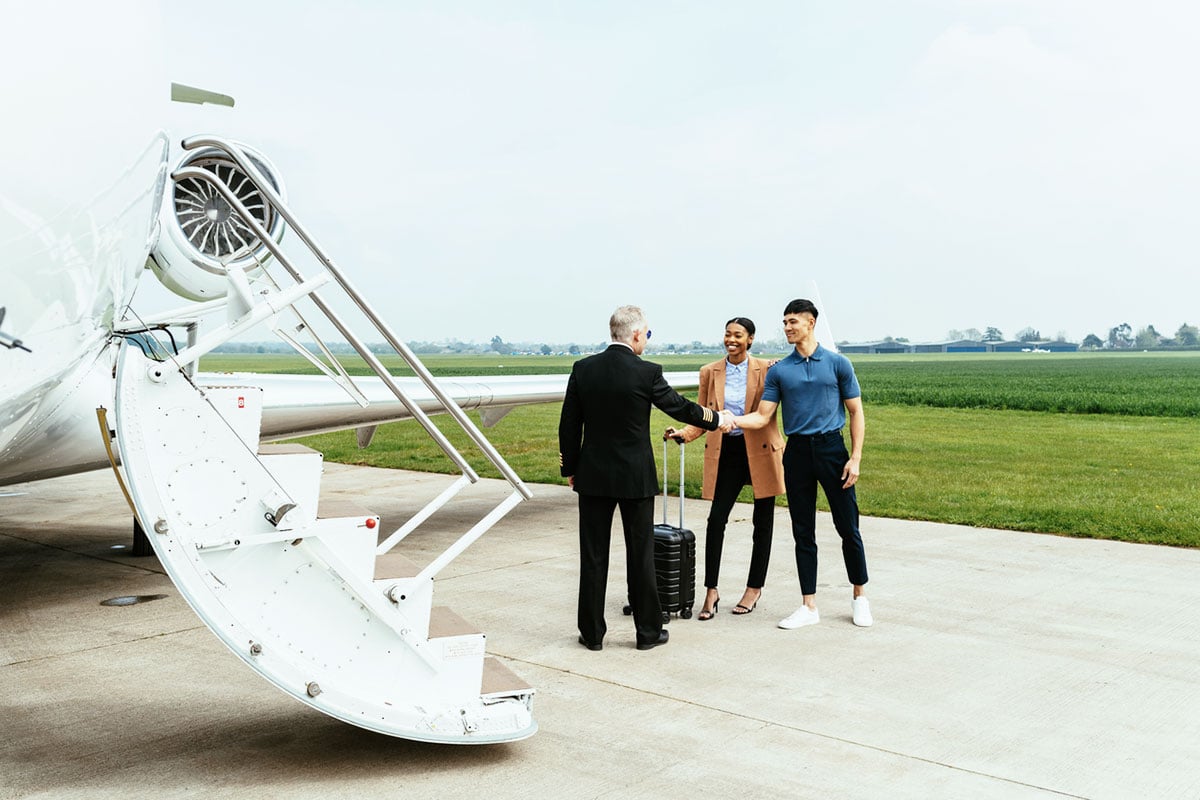 Two travelers with luggage greeted by a pilot at the stairs of a private jet on a tarmac.