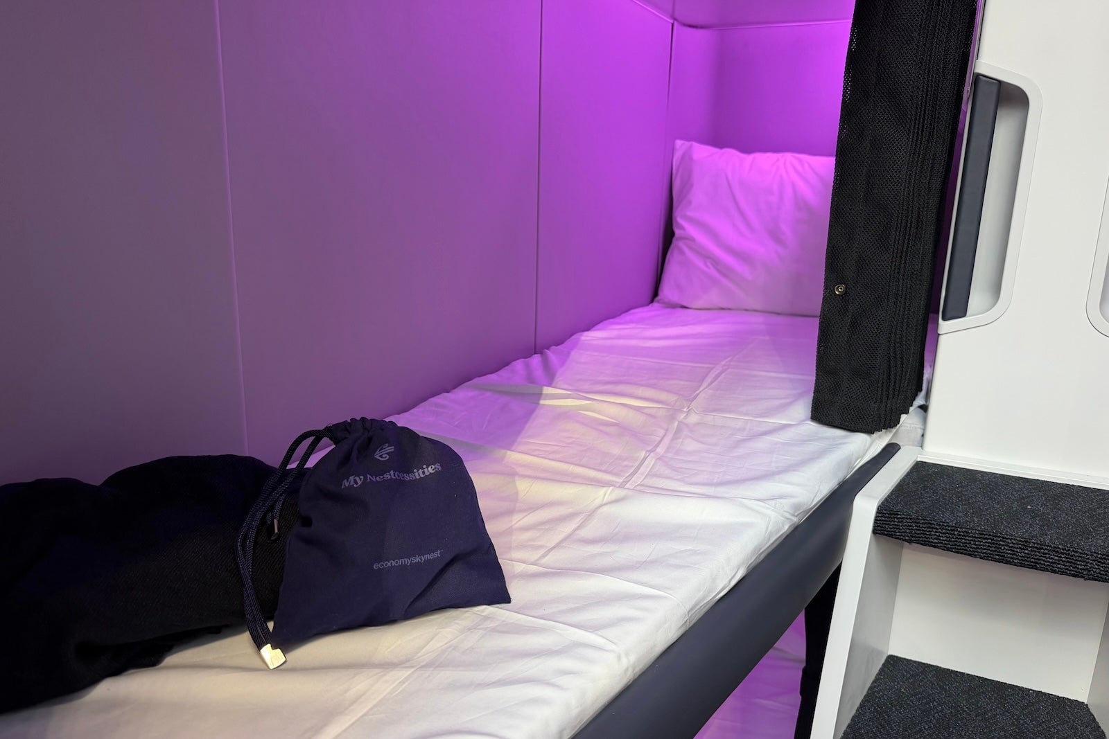 A single Air New Zealand Skynest sleep pod with purple ambient lighting, white bedding, and an amenity bag.