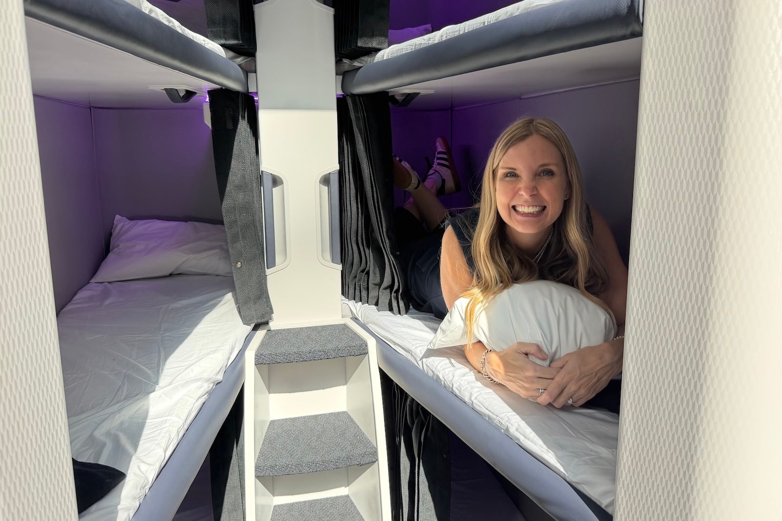 Smiling woman lying on a lower bunk in Air New Zealand's Skynest bunk bed pod, with purple lighting.
