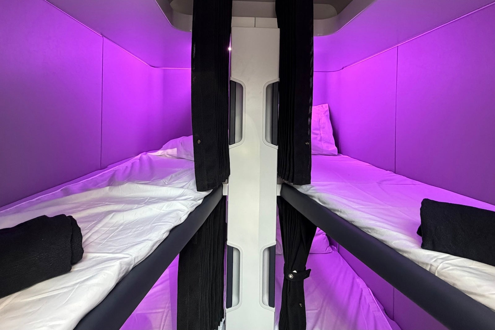 Symmetrical view of four Air New Zealand Skynest bunk pods with purple lighting, white bedding, and black curtains.